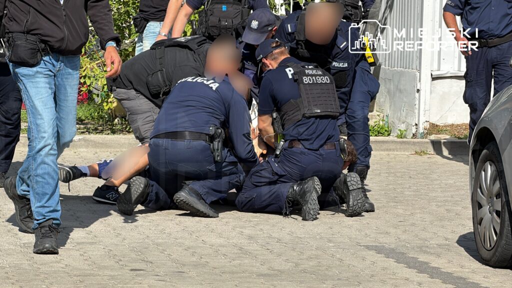Grupa funkcjonariuszy policji zatrzymuje osobę na ulicy, leżącą na ziemi, w otoczeniu innych ludzi.
