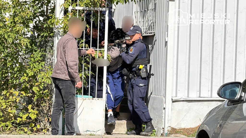 Policjant trzyma mężczyznę w niebieskich spodniach, wyprowadzając go z budynku przez bramkę, podczas interwencji.