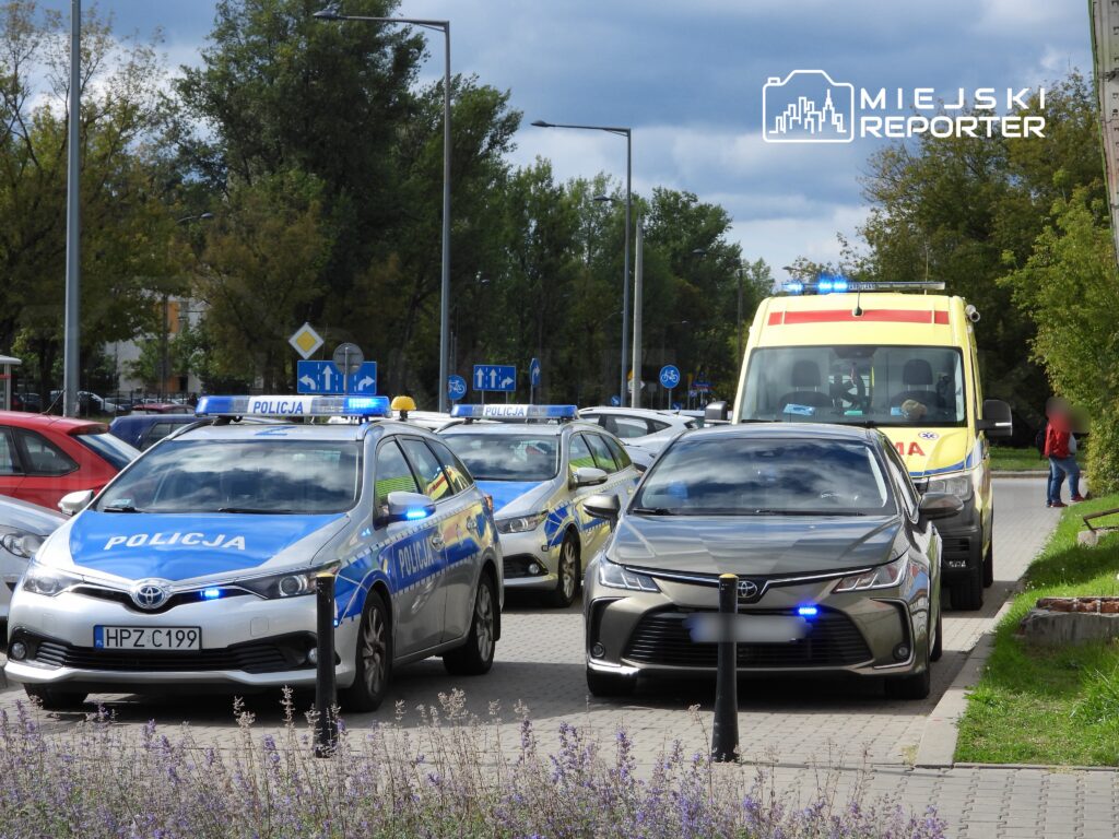 Na ulicy stoją dwa radiowozy policyjne z włączonymi sygnałami świetlnymi oraz karetka pogotowia.
