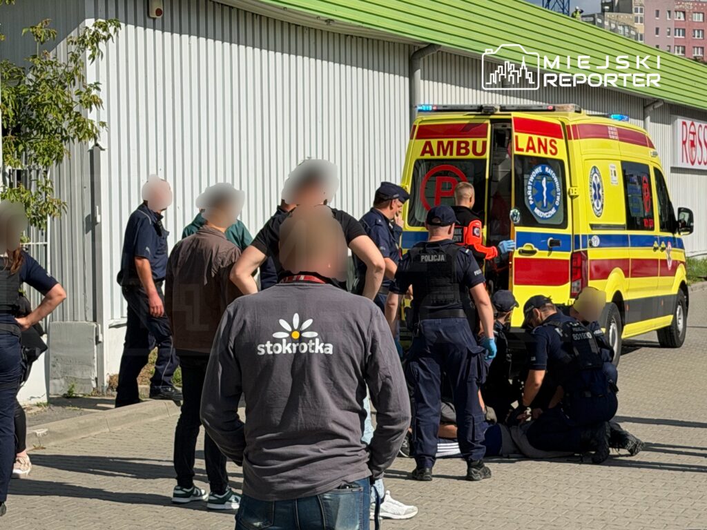 Grupa policjantów oraz ratowników medycznych udziela pomocy mężczyźnie leżącemu na ziemi obok karetki.