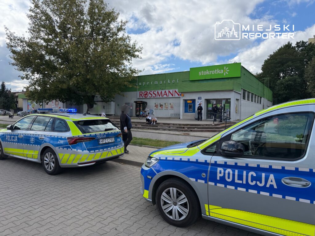 Policyjny radiowóz z włączonymi sygnałami świetlnymi stoi na chodniku obok supermarketu, a w tle widać grupę osób.