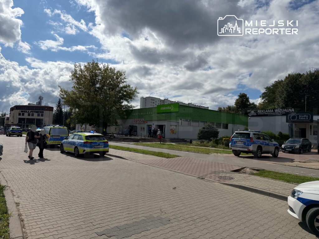 Grupa policjantów rozmawia z przechodniami na chodniku obok supermarketu, w tle widać radiowozy.