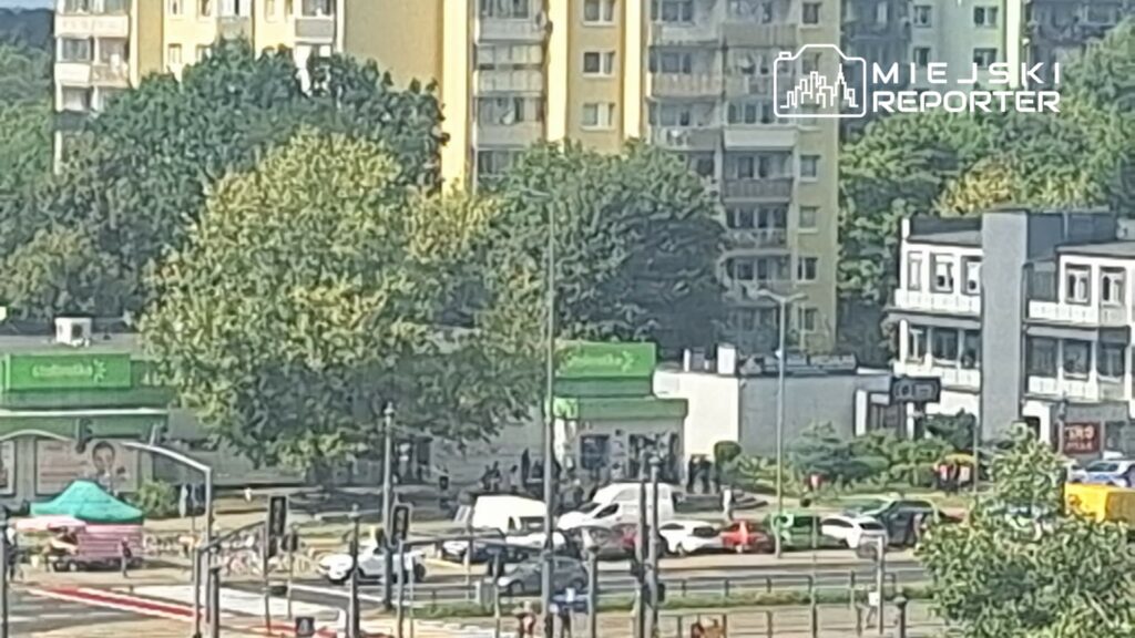 Grupa ludzi gromadzi się przed zielonym sklepem w miejskim otoczeniu, obok ruchliwej ulicy i drzew.
