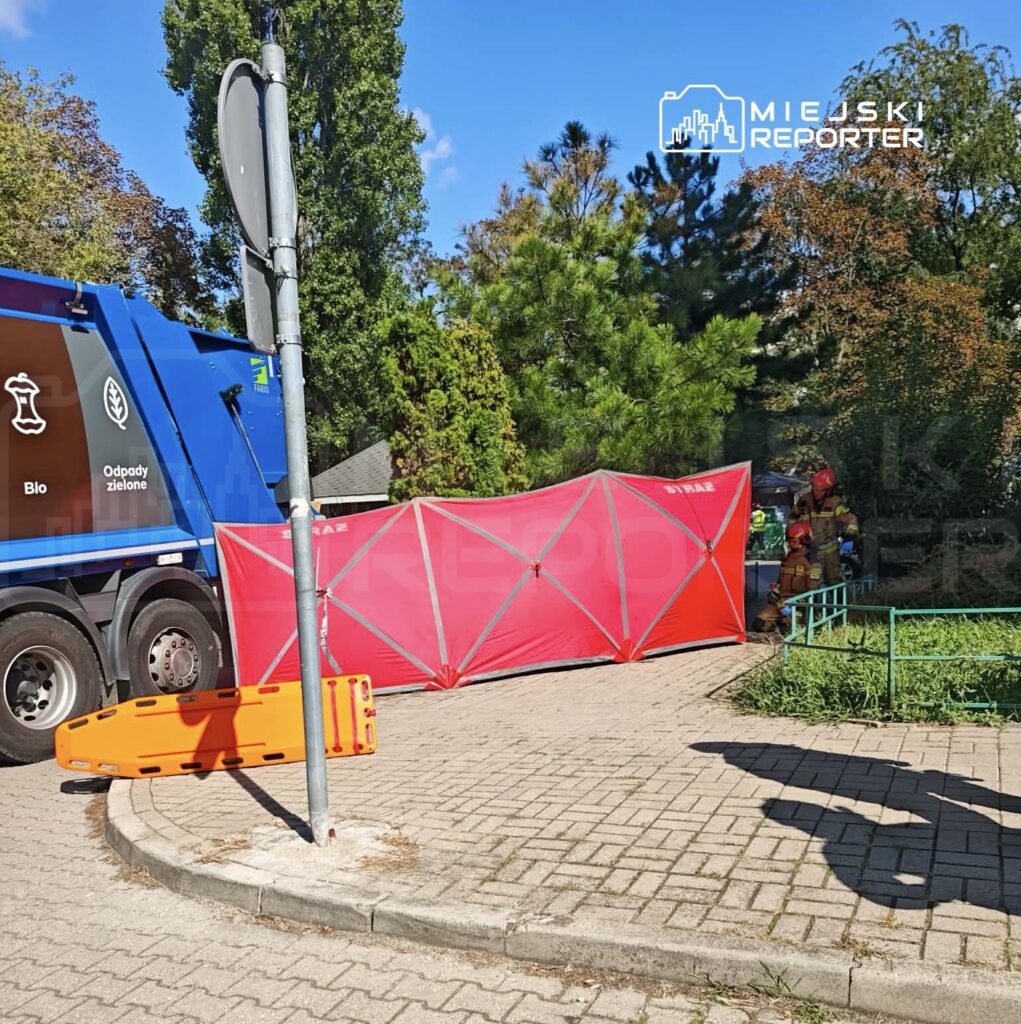 Czerwony namiot ratunkowy rozstawiony obok niebieskiego samochodu śmieciarskiego w parku, w tle strażacy przygotowują się do akcji.