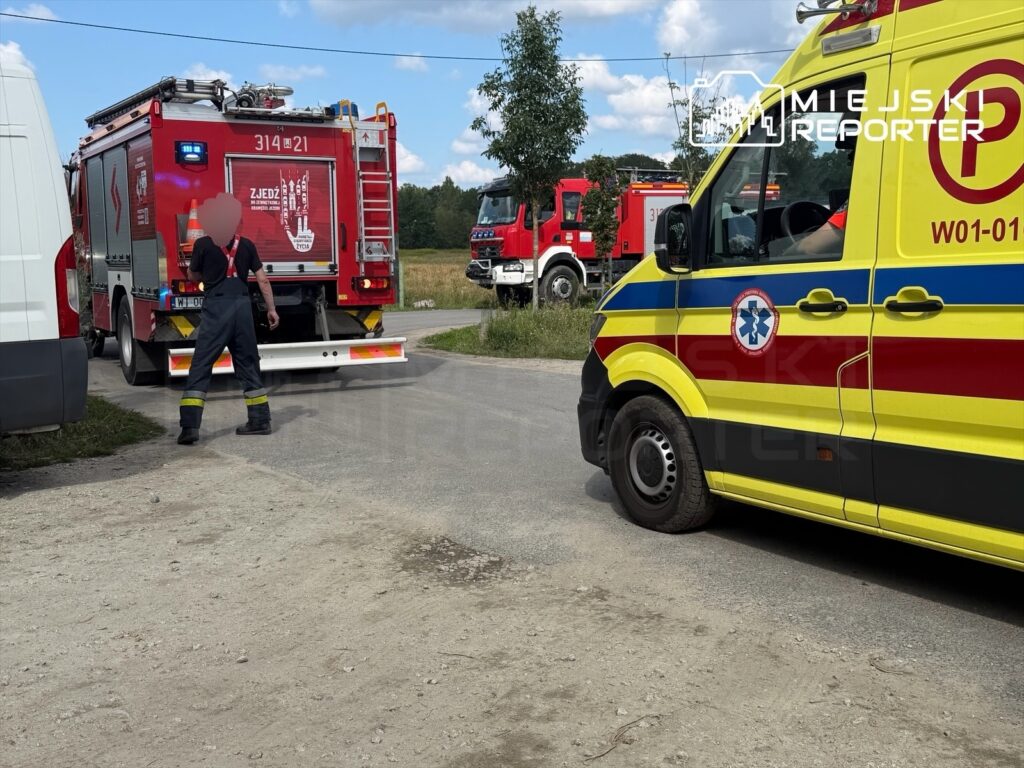 Strażak w mundurze kieruje ruchem obok wozu strażackiego i karetki na drodze w otoczeniu polnych terenów.