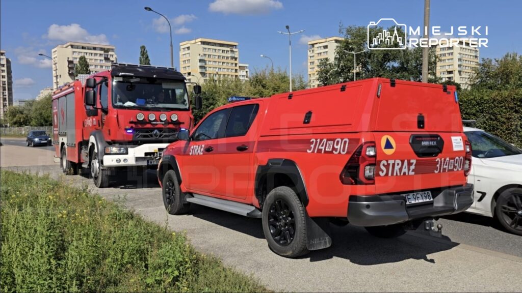 Czerwony wóz strażacki i pickup z napisem "STRAŻ" zatrzymane na drodze, w tle bloki mieszkalne i błękitne niebo.