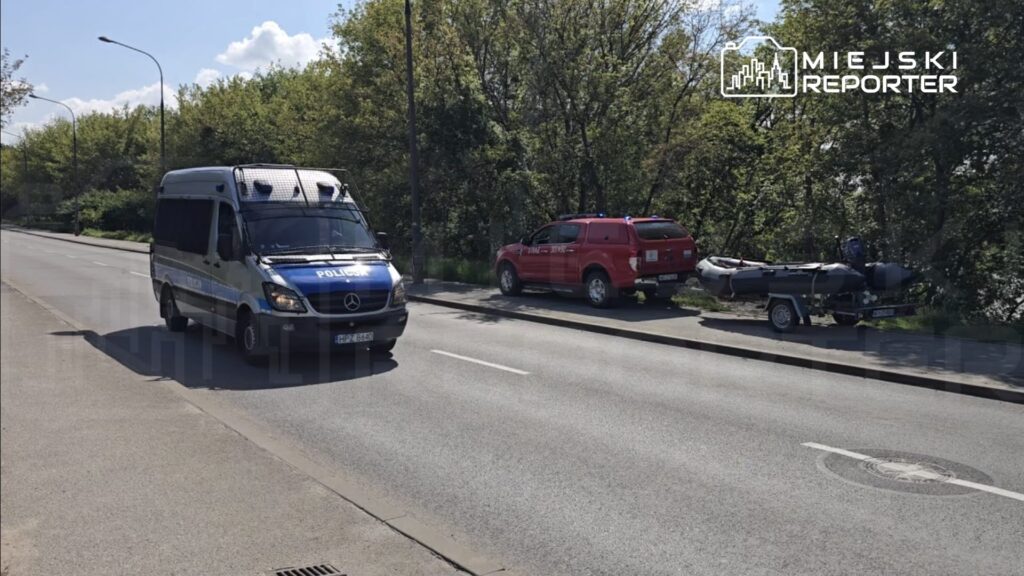 Policyjny furgon z niebieskimi sygnalizatorami porusza się po drodze, obok zaparkowany czerwony samochód terenowy oraz przyczepa z łodzią.