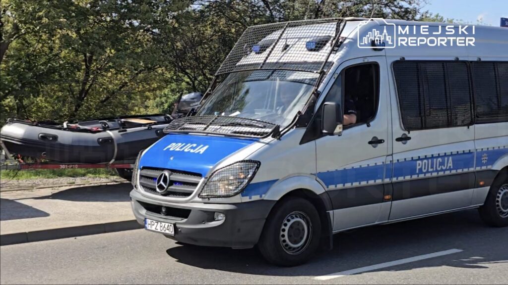 Policyjny samochód dostawczy z napisem "POLICJA" zatrzymany na drodze, obok łódź ratunkowa.