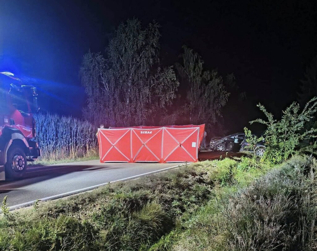 Na drodze nocą stoi czerwony namiot ratunkowy, obok uszkodzony samochód, a w tle widoczne są zboża.
