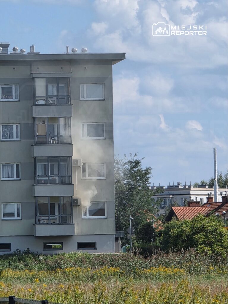 Z okna bloku wydobywa się dym, w tle widać zieloną roślinność i inne budynki.