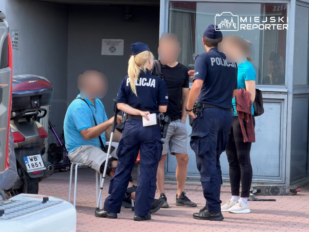 Mężczyzna na krześle rozmawia z policjantami w pobliżu budynku, obok stoi kilka osób.