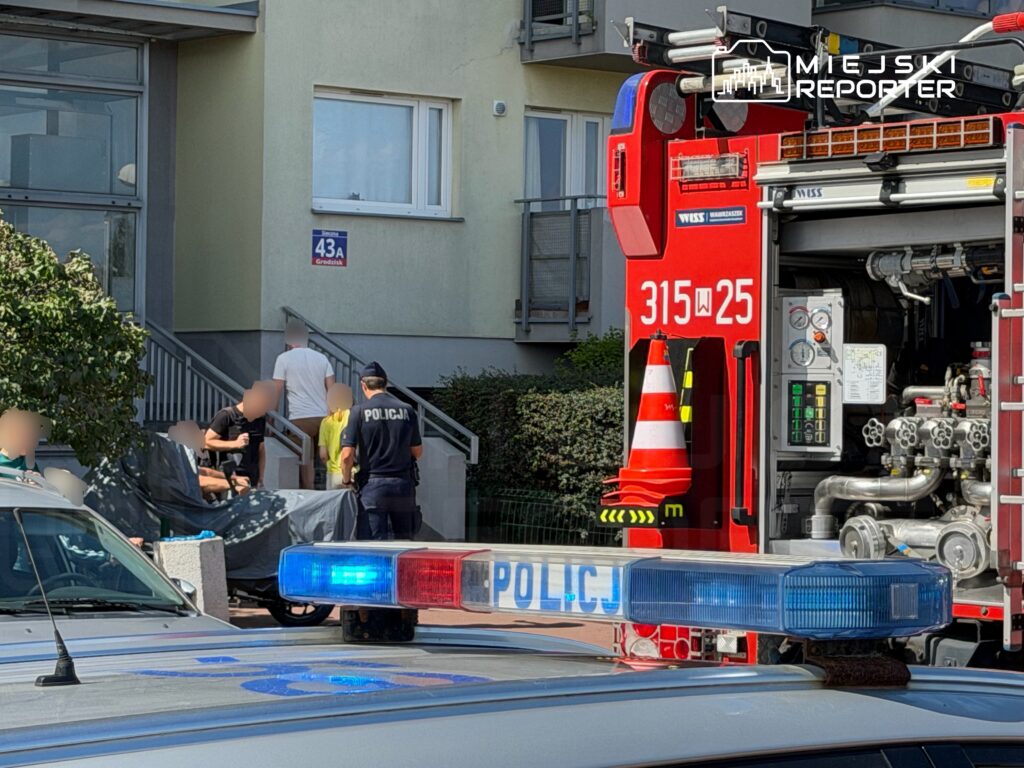 Grupa osób wchodzi po schodach budynku mieszkalnego, obok stoi wóz strażacki i radiowóz policji.