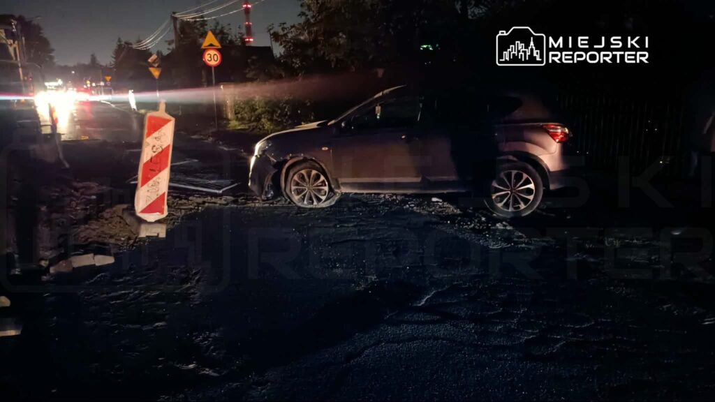 Samochód osobowy zaparkowany na uszkodzonej nawierzchni drogi, obok znak drogowy z ograniczeniem prędkości.