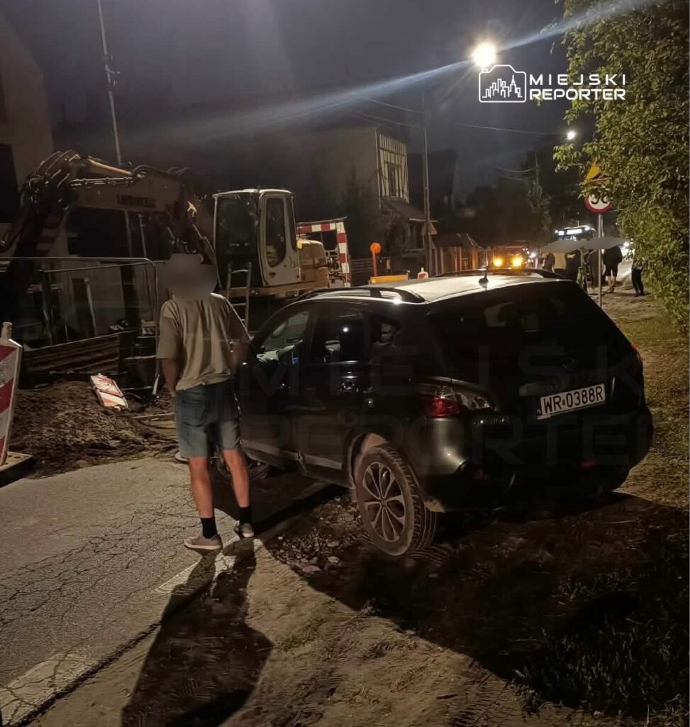 Mężczyzna w szortach stoi obok zaparkowanego samochodu na drodze, w tle widoczna koparka i oświetlenie nocne.
