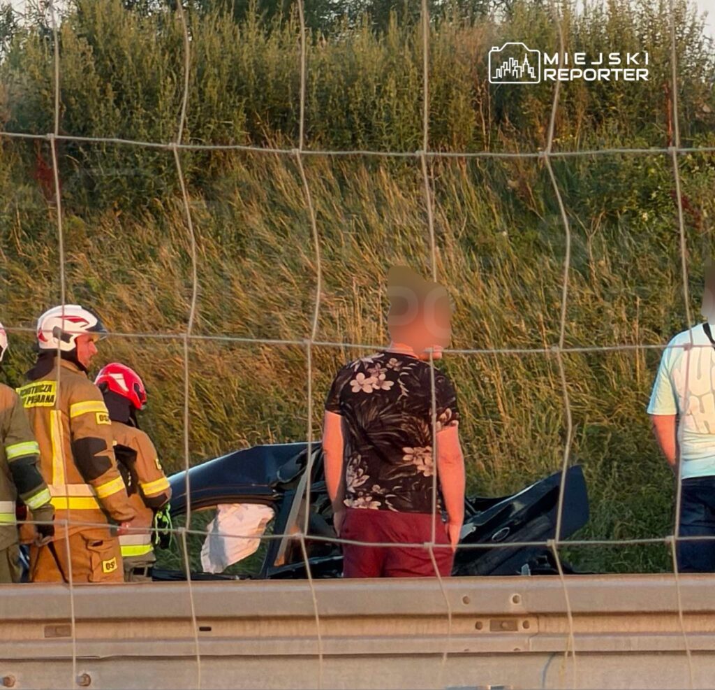 Grupa osób w pobliżu uszkodzonego samochodu na poboczu drogi, w otoczeniu wysokiej trawy.