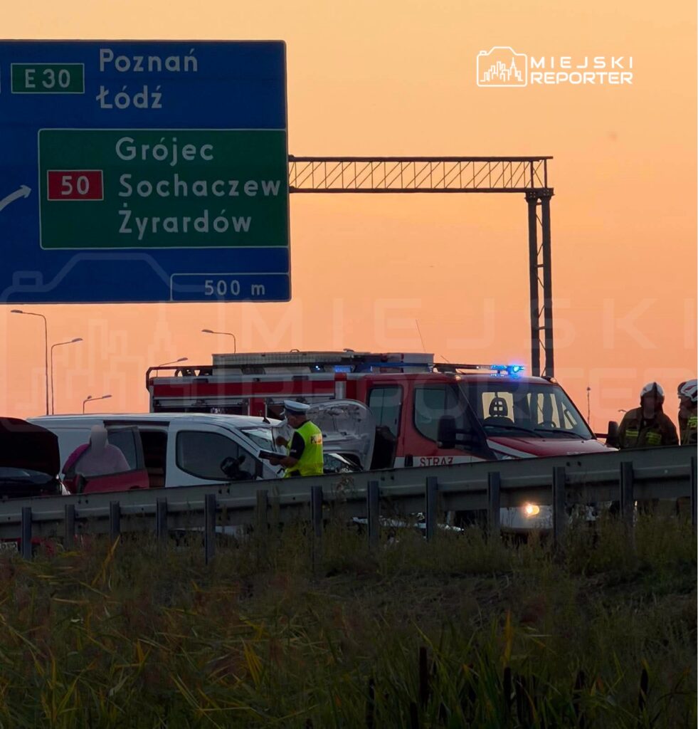 Na drodze stoi kilka pojazdów, obok nich pracują strażacy i policjanci, a w tle widoczny jest znak drogowy.
