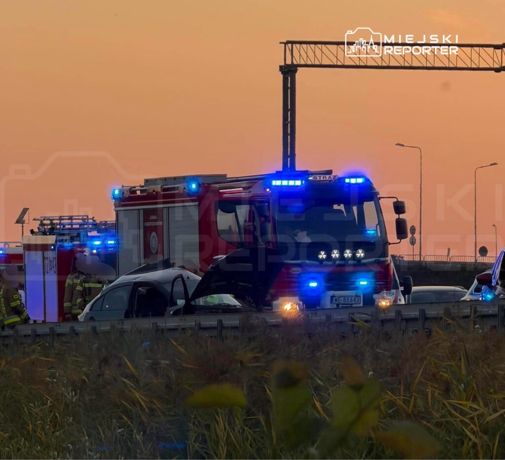 Samochód strażacki z włączonymi sygnalizatorami świetlnymi stoi obok uszkodzonego pojazdu na drodze w zachodzącym słońcu.