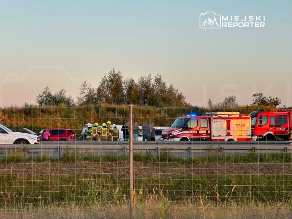 Grupa strażaków w kaskach pomaga poszkodowanym przy wypadku drogowym na autostradzie, otoczona przez pojazdy.