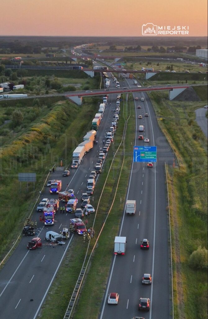 Na drodze stoi zator spowodowany wypadkiem, w którym uczestniczy kilka pojazdów, a w tle widać karetki i straż pożarną.