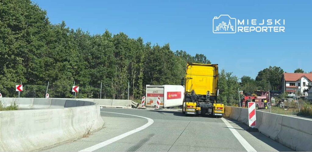 Żółty ciężarówka skręca na wąskiej drodze, obok znajdują się znaki ostrzegawcze i zieleń drzew.