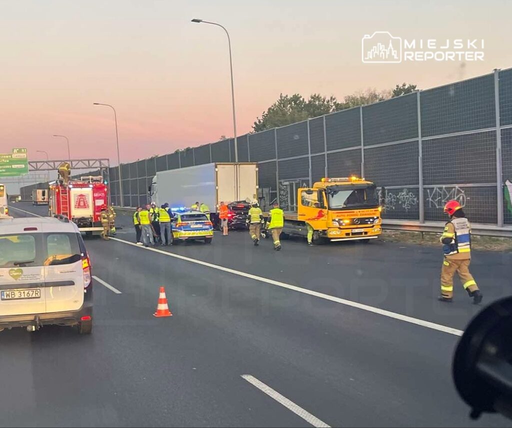 Grupa ratowników i policjantów zabezpiecza miejsce wypadku na autostradzie, obok ciężarówki i wozu strażackiego.