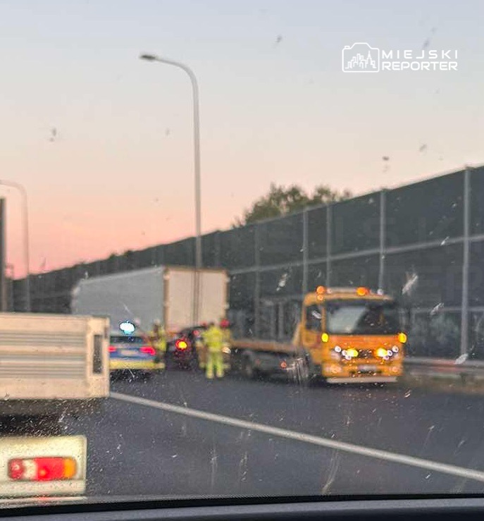Kierowcy zatrzymani w korku na drodze, obok ciężarówki i policyjnych pojazdów, w tle zachód słońca.