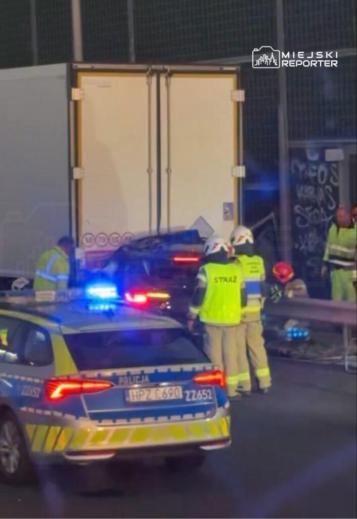 Strażacy w kamizelkach odblaskowych pracują przy wypadku drogowym, obok policyjnego samochodu z włączonymi sygnałami świetlnymi.