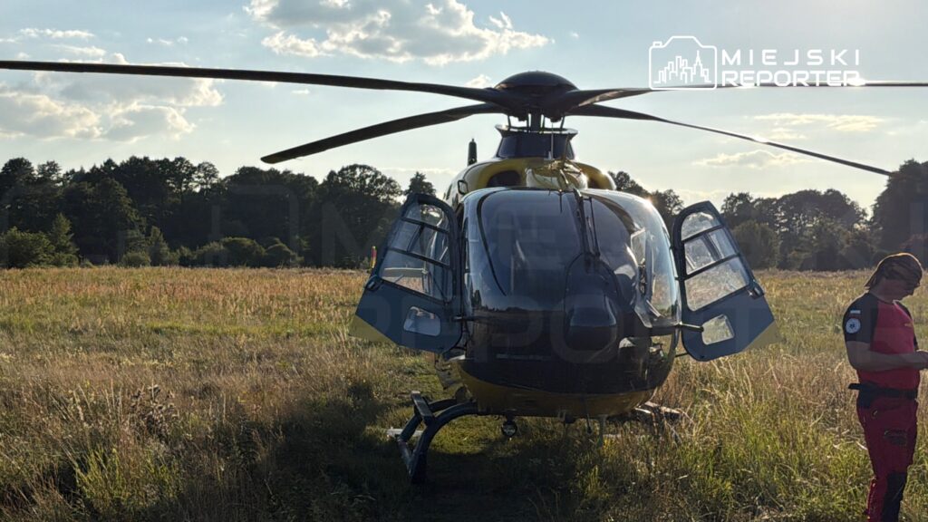 Helikopter ratunkowy ląduje na otwartej przestrzeni, obok stoi ratownik w czerwonym stroju, sprawdzający sprzęt.