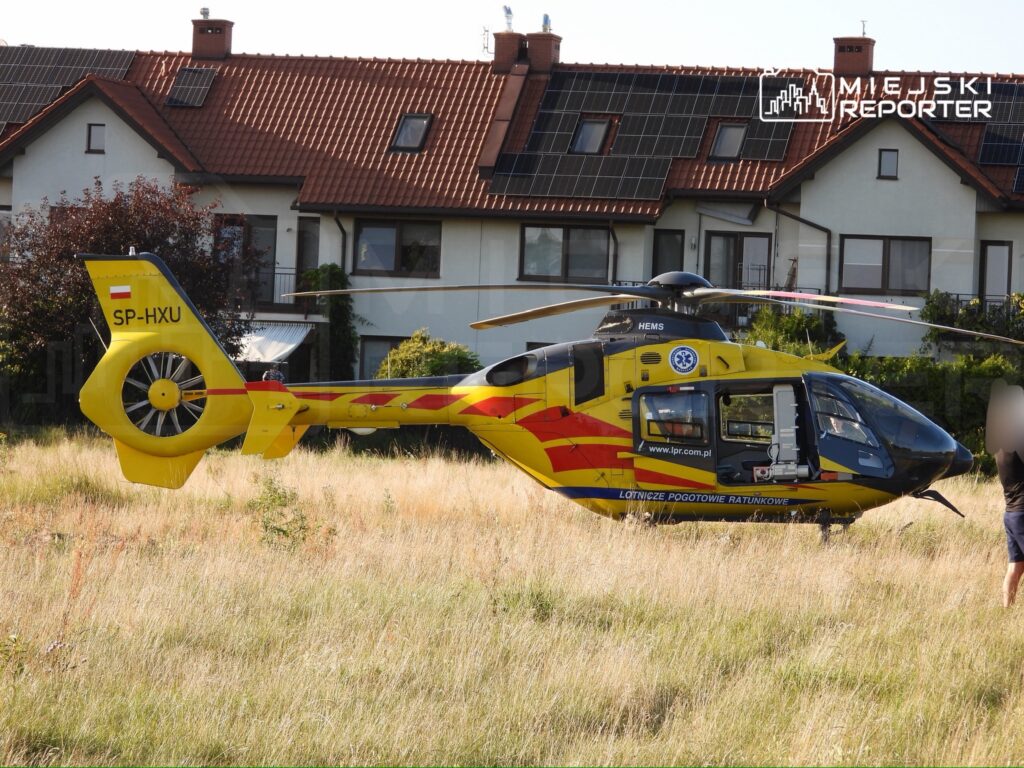 Śmigłowiec ratunkowy lądował na trawie obok budynków mieszkalnych, z widocznym logo HEMS i napisem "LOTNICZE POGOTOWIE RATUNKOWE".