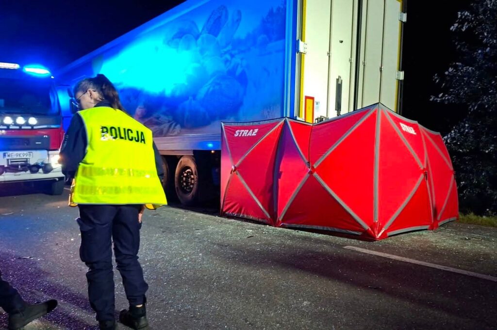 Policjant w kamizelce odblaskowej bada miejsce wypadku drogowym obok ciężarówki z niebieskimi światłami.