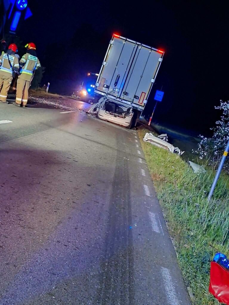 W nocy na drodze stoi ciężarówka, a obok niej grupa strażaków bada miejsce wypadku. W tle widać sygnalizację świetlną.