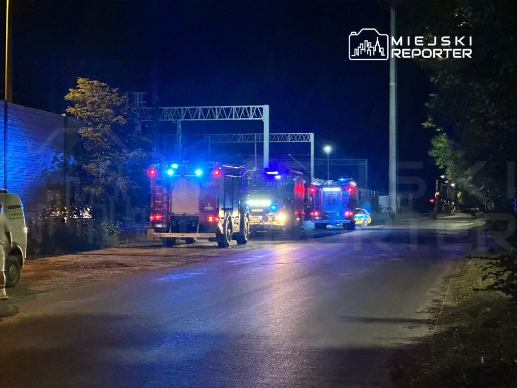 W nocy na drodze zatrzymane są pojazdy straży pożarnej i policji, emitujące niebieskie światła.