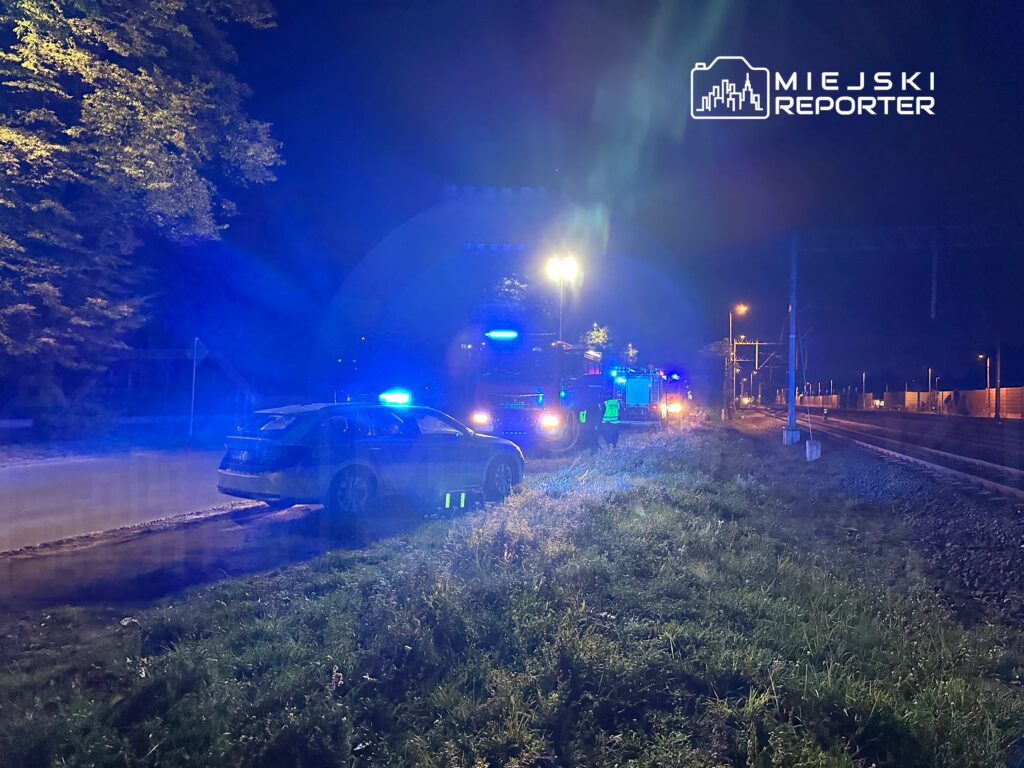Nocna akcja służb ratunkowych przy torach kolejowych, z widocznymi pojazdami z niebieskimi światłami.