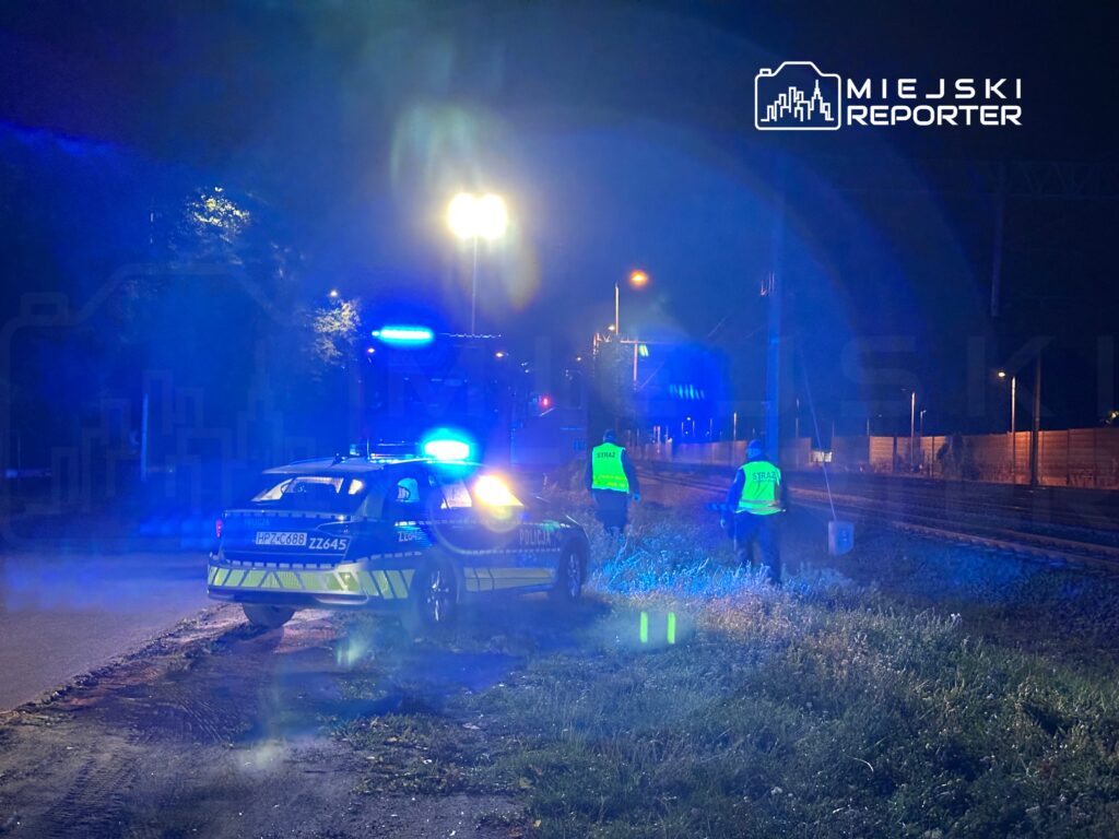 Policjanci i strażacy w odblaskowych kamizelkach sprawdzają teren przy torach kolejowych w nocy, otoczeni niebieskim światłem.