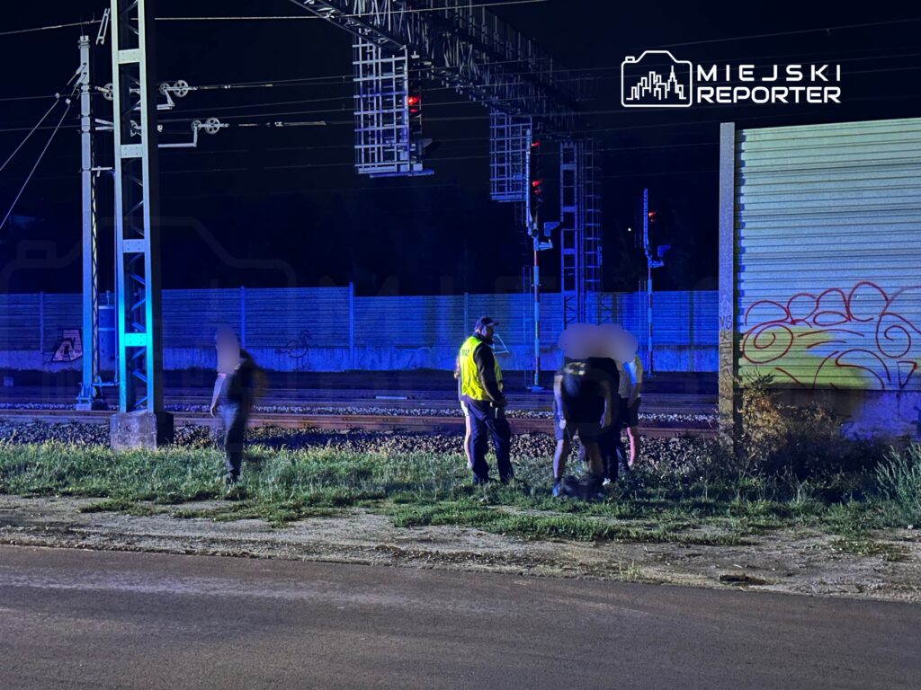 Grupa osób w odzieży roboczej bada teren przy torach kolejowych w nocy, w otoczeniu słupów trakcyjnych.