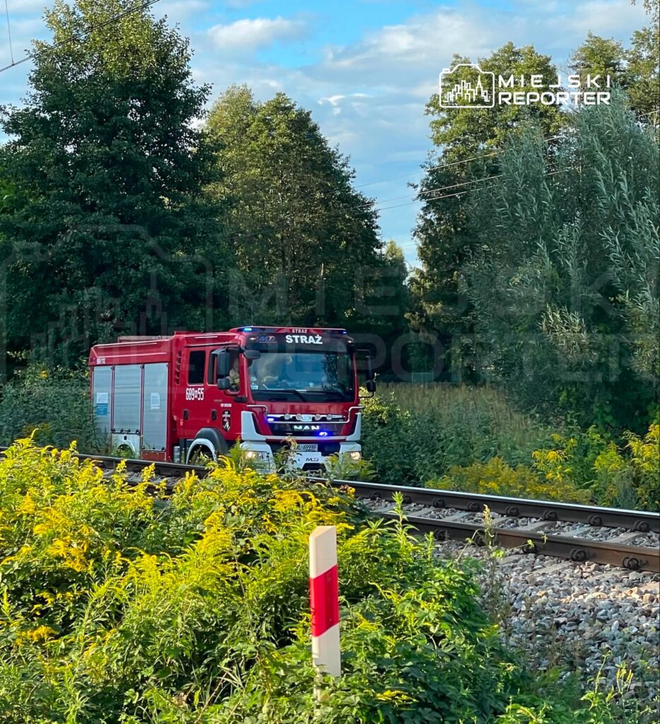 Czerwony wóz strażacki MAN przejeżdża obok torów kolejowych, otoczony zielenią i krzewami.