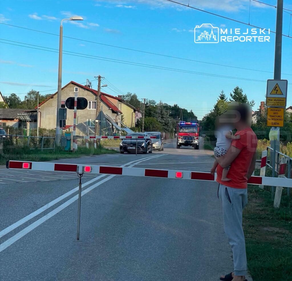 Mężczyzna w czerwonej koszulce trzyma dziecko, stojąc przy opuszczonym szlabanem drogowym. W tle widoczny wóz strażacki.