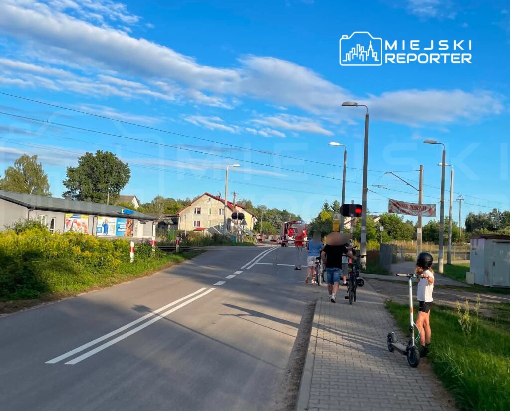 Grupa osób czeka na przejeżdżający pociąg przy przejeździe kolejowym, obok drogi i budynków mieszkalnych.
