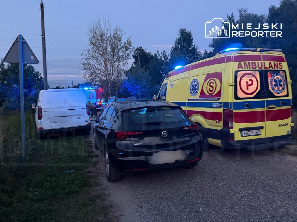 Karetka pogotowia z włączonymi sygnałami świetlnymi stoi obok dwóch zaparkowanych samochodów na wąskiej drodze.