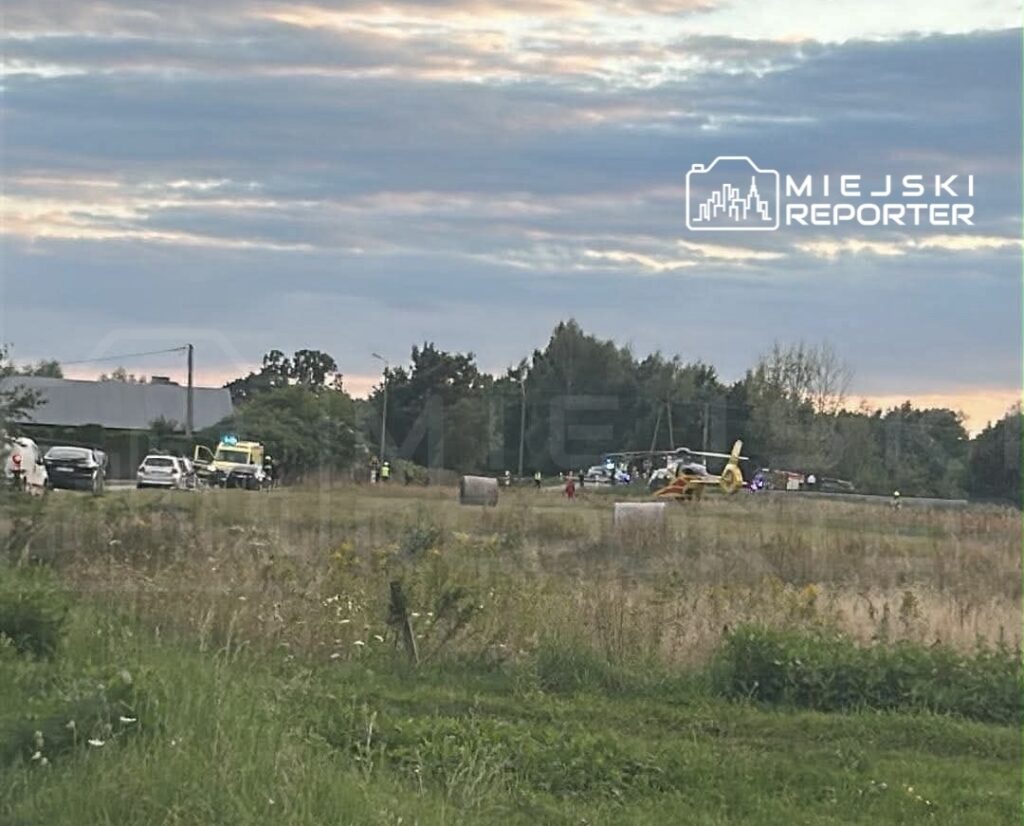 Na polu lądował helikopter ratunkowy, a obok stoją pojazdy służb ratunkowych oraz grupy ludzi.