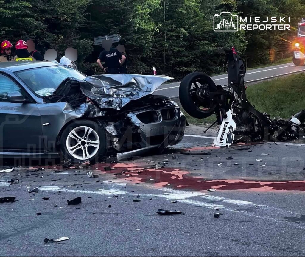 W wyniku wypadku drogowego uszkodzone auto osobowe z rozbitym przodem oraz przewrócony motocykl, w otoczeniu służb ratunkowych.