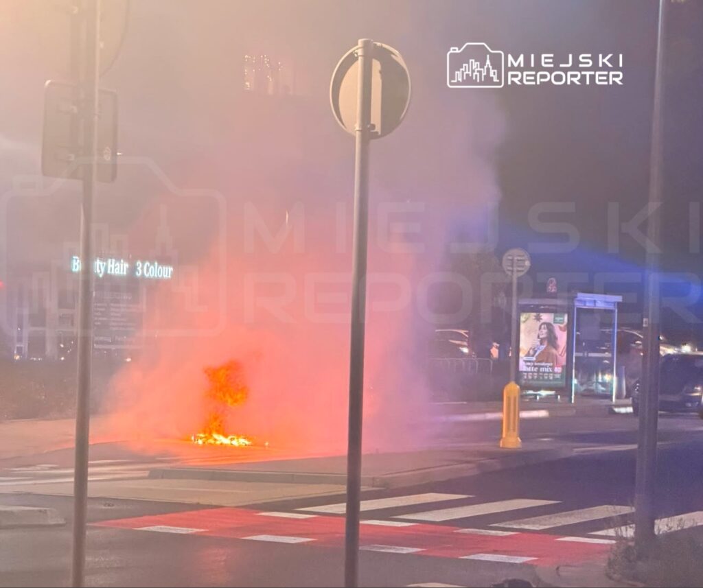 Na chodniku unosi się dym z płonącego materiału, w tle widoczne znaki drogowe oraz przystanek autobusowy.