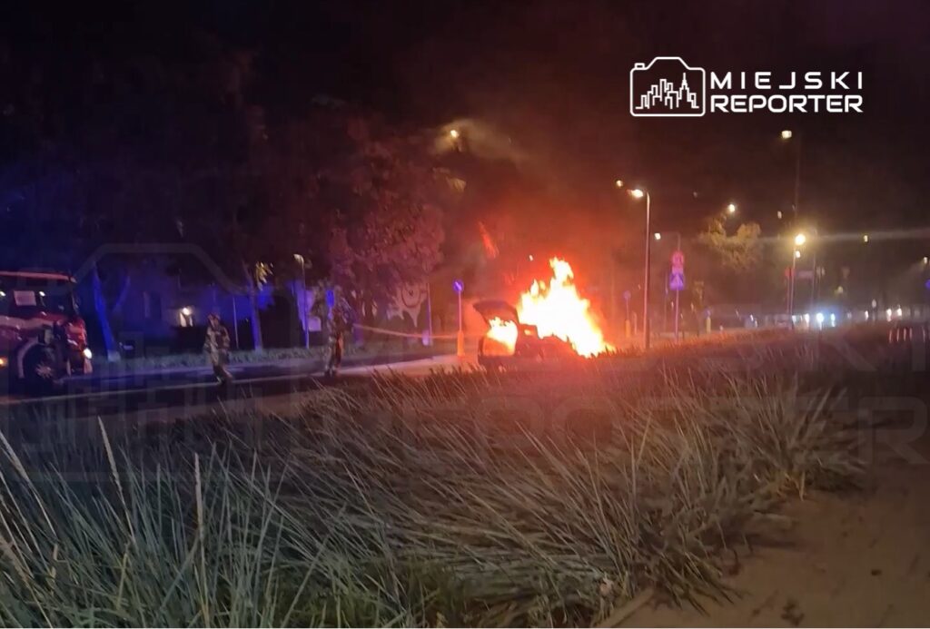 Pojazd w płomieniach na ulicy, obok strażacy w mundurach gaszący ogień, nocna sceneria z oświetleniem ulicznym.