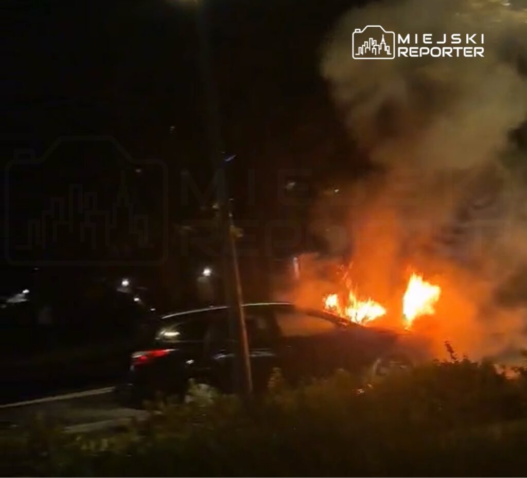 Samochód pali się na poboczu drogi, otoczony przez dym i płomienie, w ciemnej scenerii nocnej.