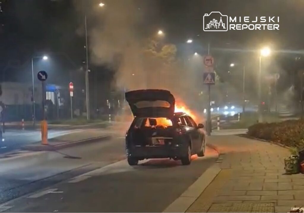 Samochód zaparkowany na ulicy płonie, wydobywając dym z otwartego bagażnika, w nocy w miejskim otoczeniu.