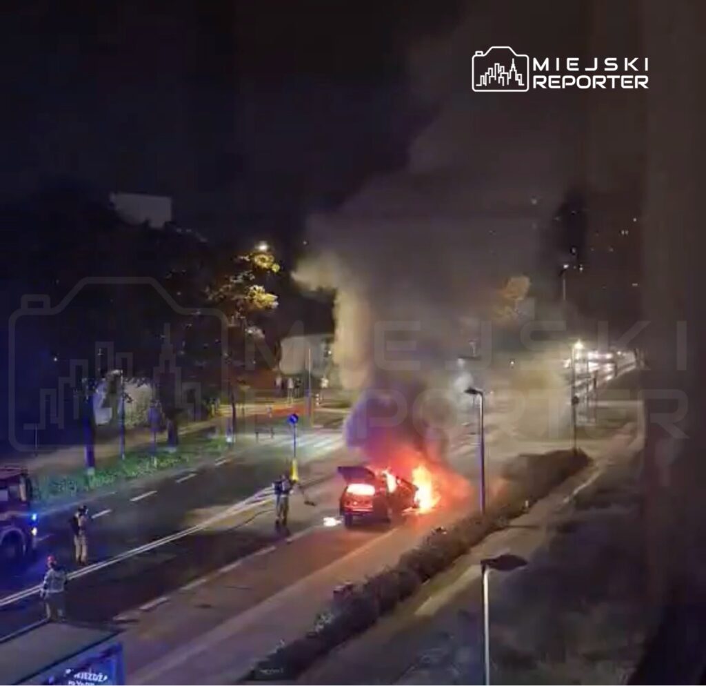 Samochód płonący na drodze, wokół strażacy i gapie obserwujący zdarzenie w nocy.