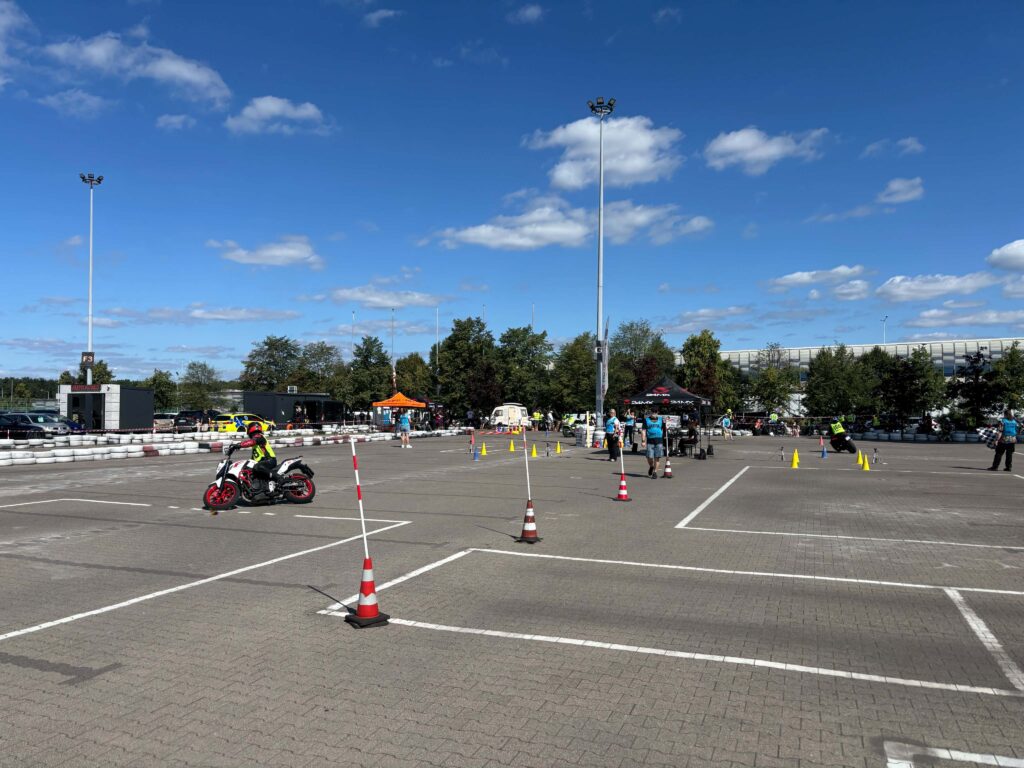 Motocyklista w kasku i kamizelce, pokonujący tor przeszkód na parkingu, otoczony przez uczestników i organizatorów wydarzenia.