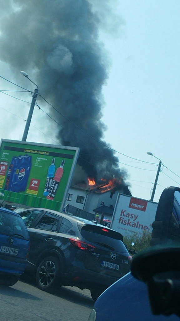 Czarny dym unoszący się z budynku, w tle widoczne samochody stojące w korku oraz billboardy reklamowe.