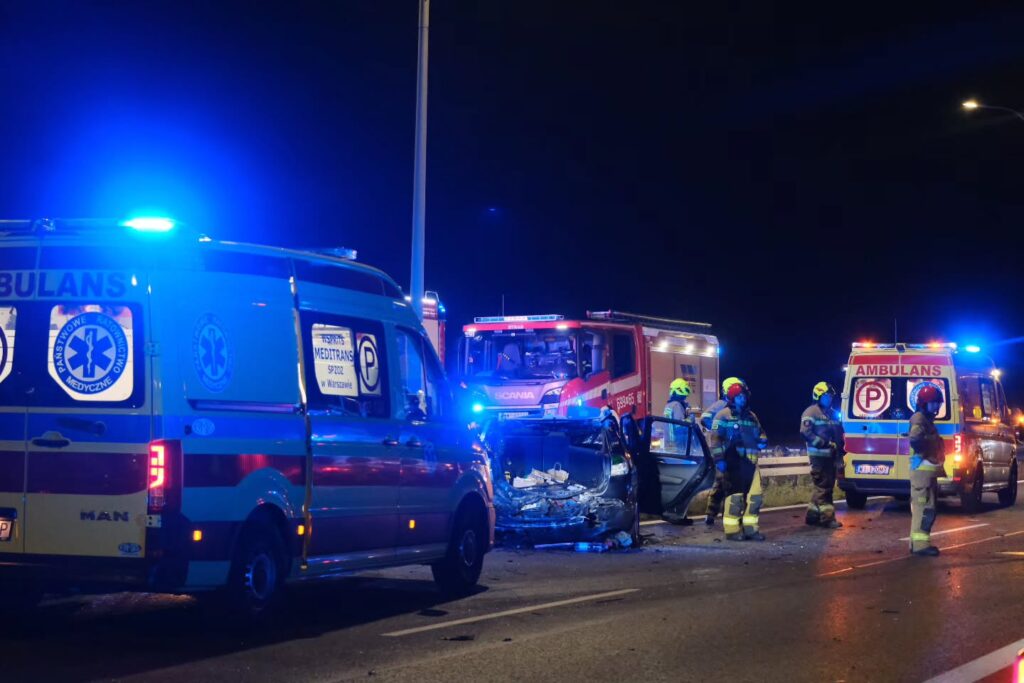 Zespół ratowników medycznych i strażaków pracuje przy rozbitym samochodzie na drodze w nocy.