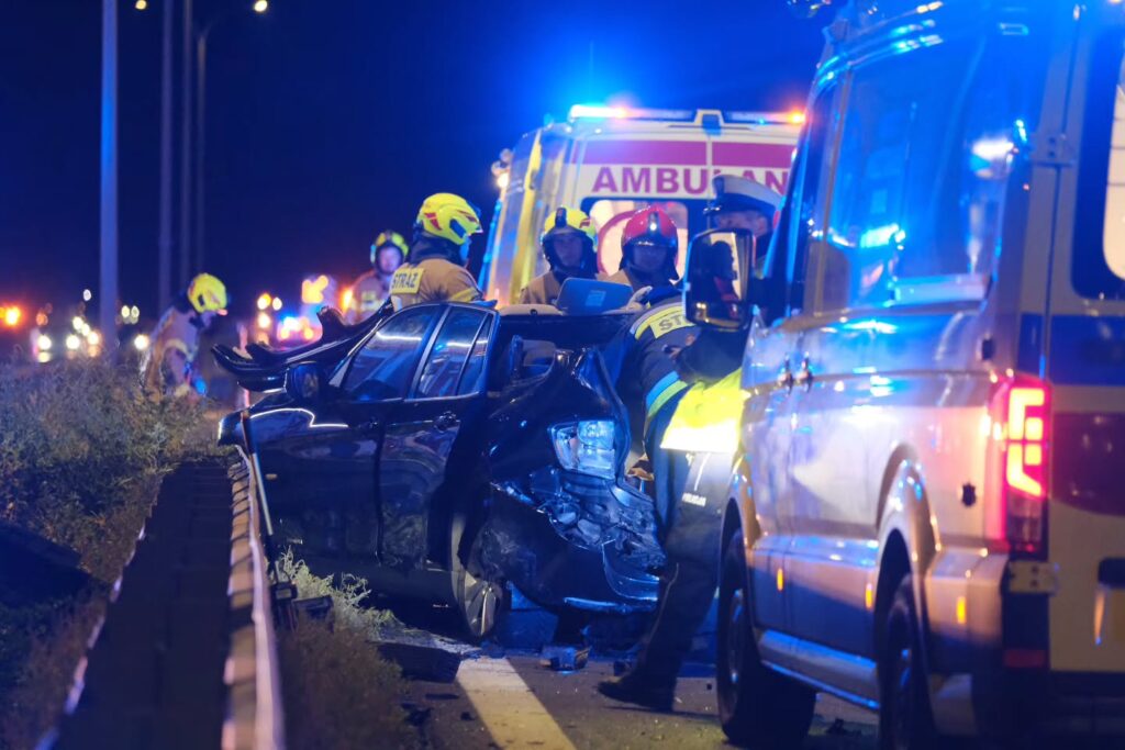 Ratownicy medyczni pracują przy rozbitym czarnym samochodzie osobowym na nocnej drodze, otoczeni przez pojazdy służb ratunkowych.